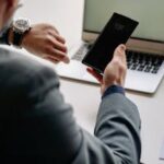 Man in office using smartphone and laptop, showing time management.